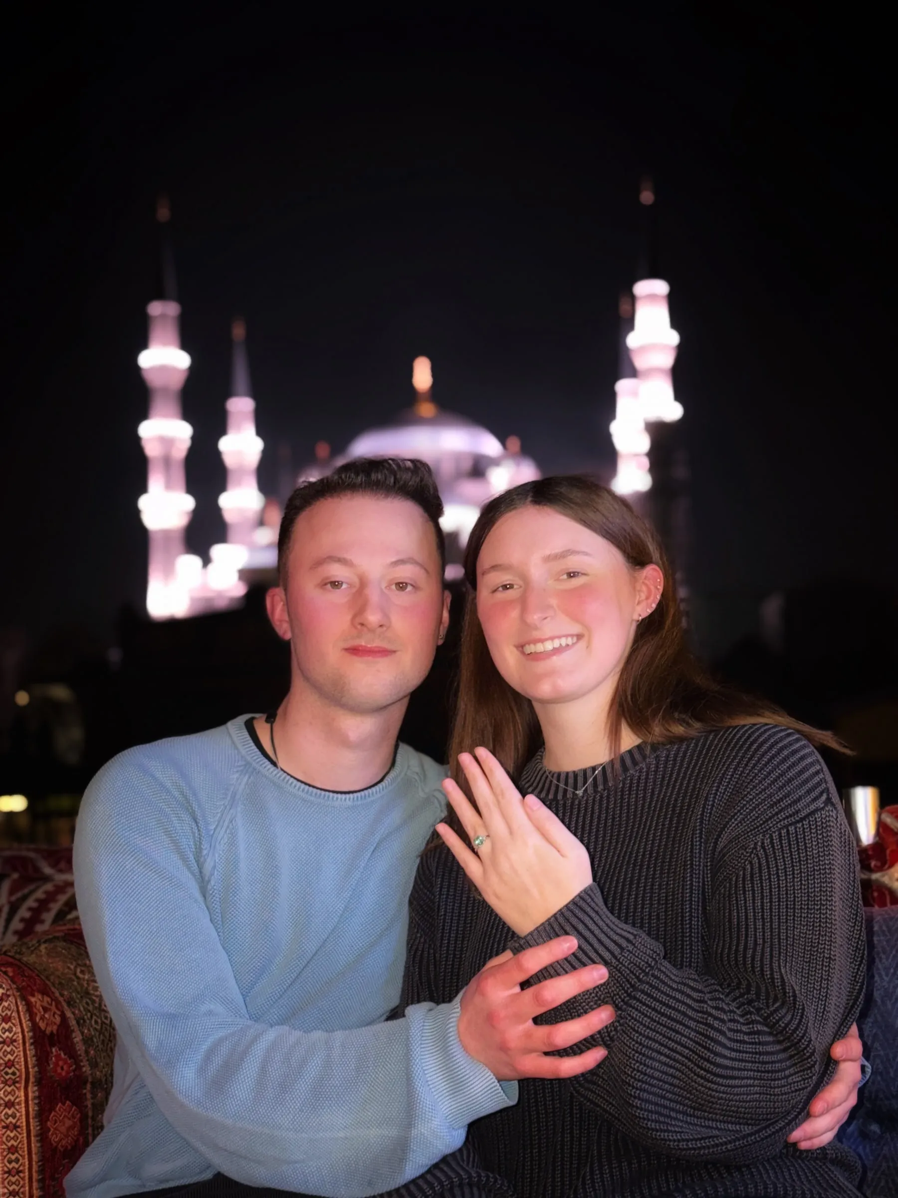 Sailor and Adilhan at night in front of a lit-up mosque. She is showing her engagement ring.